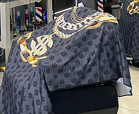 Capa negra de la barbería estampada con signos de pesos y cadenas doradas, sobre un sillón