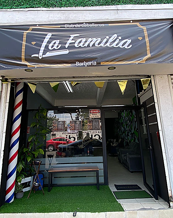 Fachada de La Familia Barbería de día: lona negra con marco dorado que dice Club de caballeros, La Familia, Barbería; poste de barbero rayado, banca de madera y pasto en la entrada