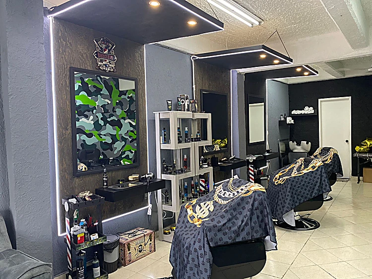 Interior de la barbería: espejo con camuflaje verde y lima pintado a mano, repisas con productos, luces de tira blanca y sillones cubiertos con capas negras con cadenas doradas